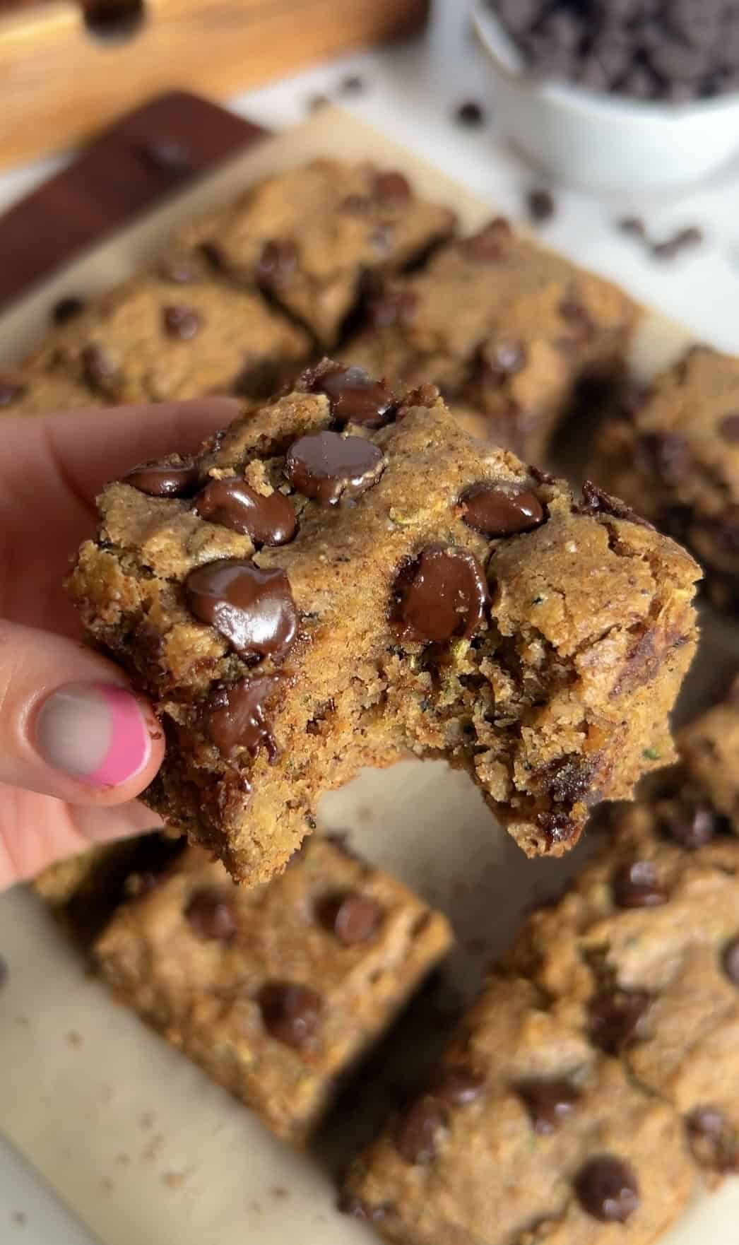 Hand holding a soft, chewy zucchini blondie square with a bite taken out, showing gooey melted chocolate chips.