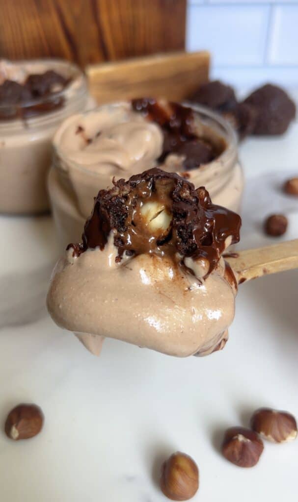 Spoonful of Ferrero Rocher cottage cheese mousse showing creamy texture and hazelnut center, surrounded by jars of chocolate mousse and scattered hazelnuts.