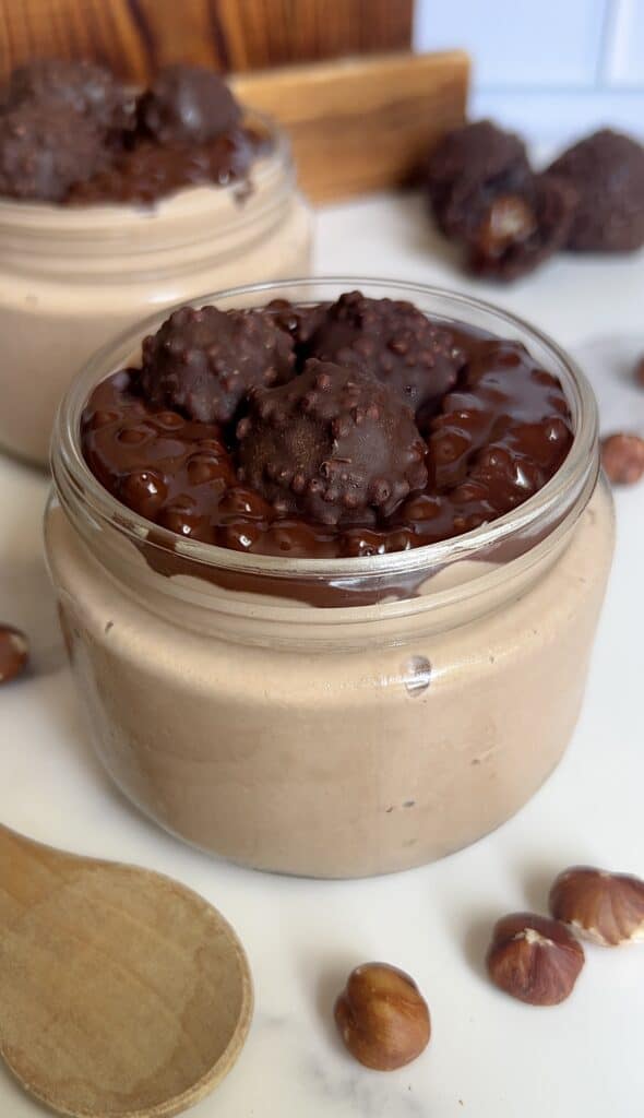 hick and creamy Ferrero Rocher cottage cheese mousse in a glass jar topped with glossy chocolate ganache and mini hazelnut truffles, surrounded by whole hazelnuts and a wooden spoon.