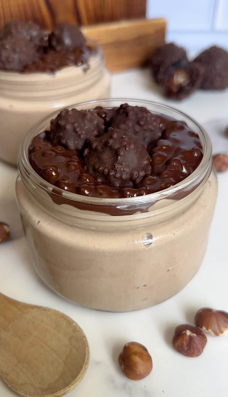 hick and creamy Ferrero Rocher cottage cheese mousse in a glass jar topped with glossy chocolate ganache and mini hazelnut truffles, surrounded by whole hazelnuts and a wooden spoon.