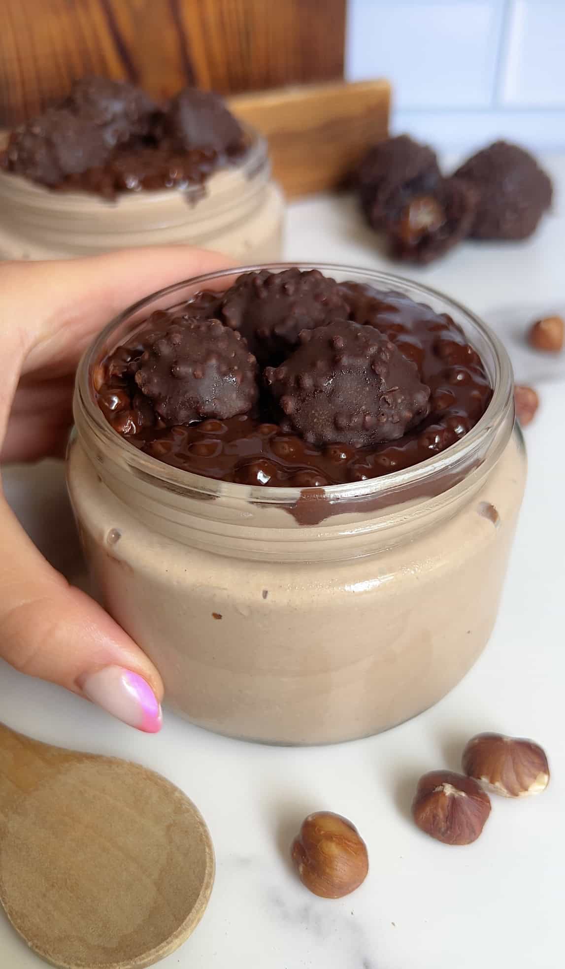 Hand holding a jar of chocolate hazelnut cottage cheese mousse topped with mini Ferrero Rocher balls and rich chocolate ganache, styled with hazelnuts on a marble surface.