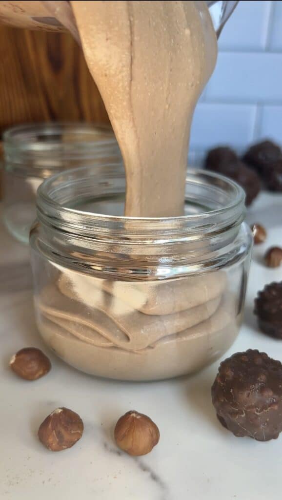 Smooth hazelnut cottage cheese mousse being poured into glass jars, surrounded by whole hazelnuts and Ferrero Rocher‑style truffles for a high‑protein dessert prep scene.