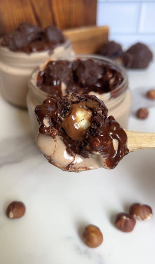 Spoonful of Ferrero Rocher cottage cheese mousse showing creamy texture and hazelnut center, surrounded by jars of chocolate mousse and scattered hazelnuts.