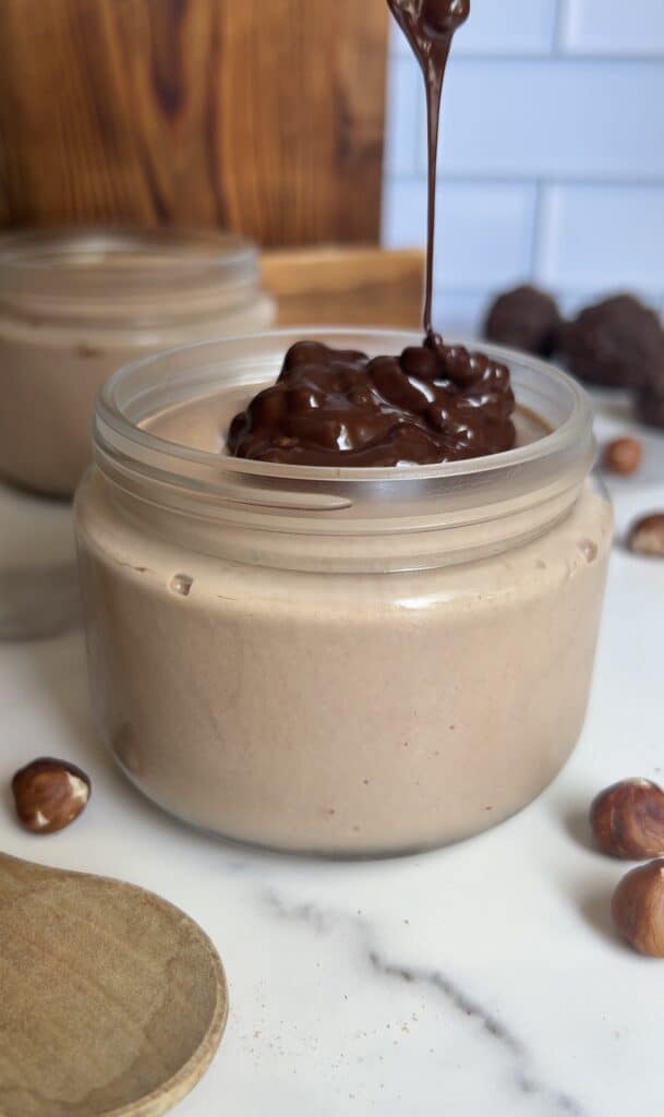 Chocolate ganache being poured over thick hazelnut cottage cheese mousse in a jar, highlighting rich texture and glossy finish with hazelnuts nearby.
