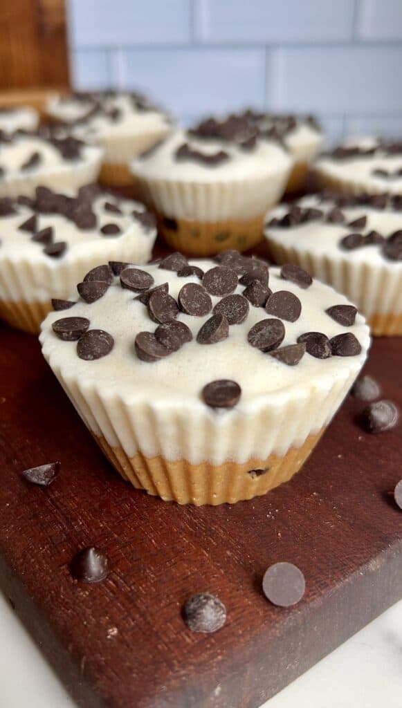 Frozen cookie dough ice cream cups with chocolate chips on top displayed on a wooden board against a tiled background