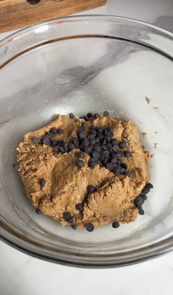 Glass mixing bowl filled with peanut butter protein cookie dough topped with mini chocolate chips on a marble countertop
