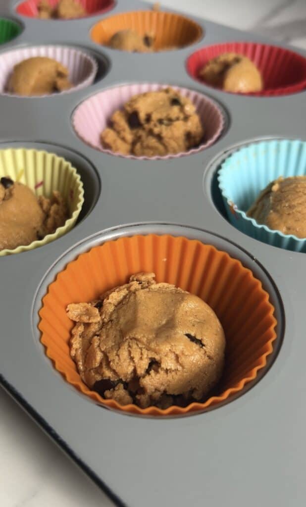 Colorful silicone muffin liners filled with scoops of peanut butter protein cookie dough in a baking tray before pressing down