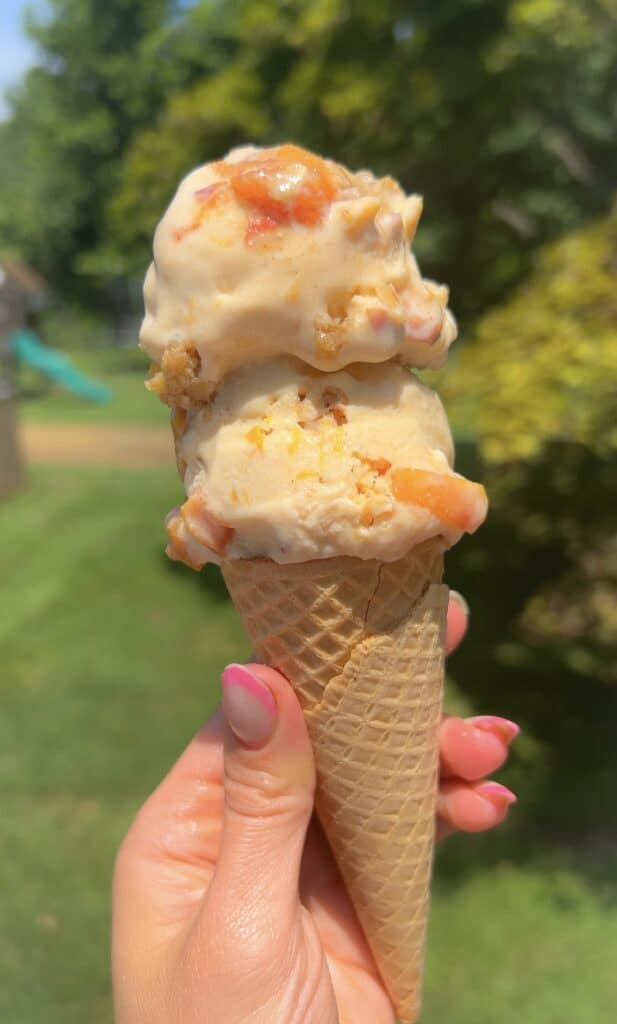 Hand holding waffle cone of high‑protein peach cobbler ice cream with peach chunks outdoors