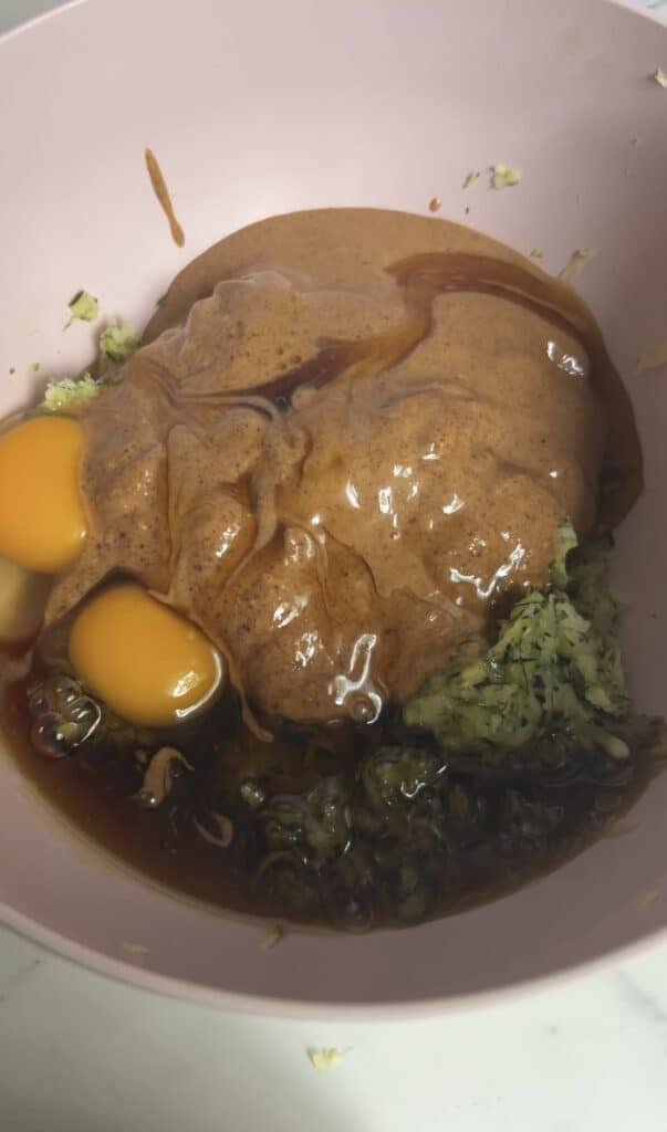 Almond butter, eggs, maple syrup, and shredded zucchini in a pink bowl before mixing for gluten‑free blondies.