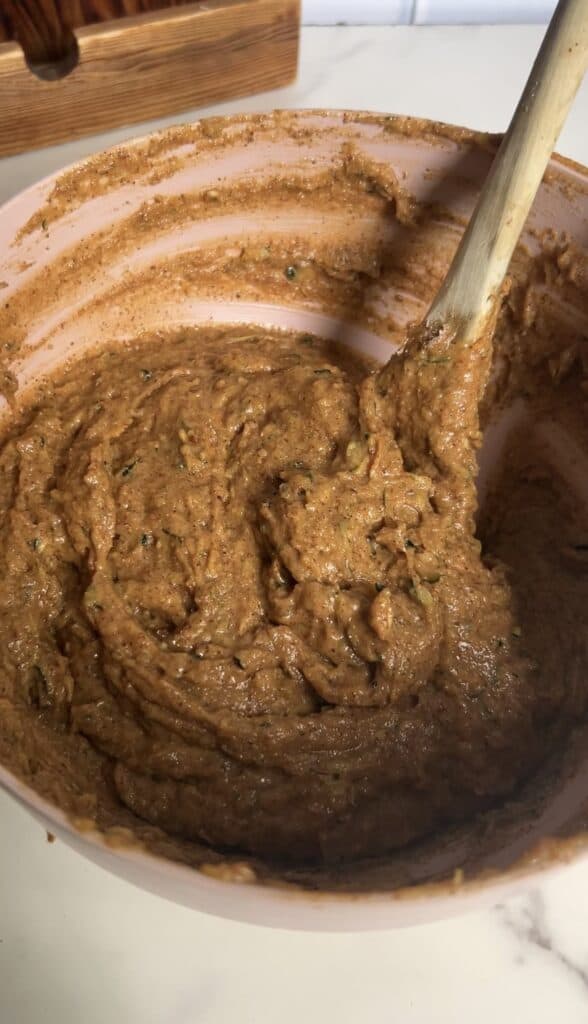 Thick, smooth zucchini blondie batter with visible green flecks and a wooden spoon in a pink bowl.