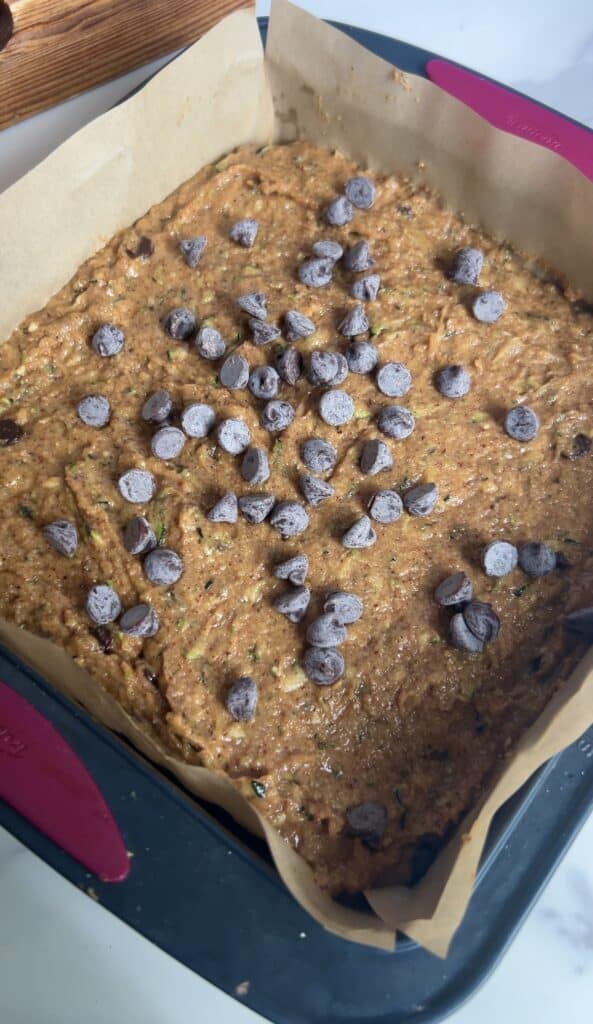 Unbaked zucchini blondie batter spread evenly in a parchment‑lined pan topped with chocolate chips