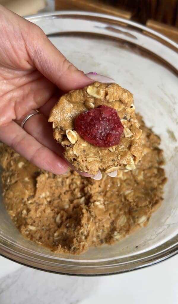 Hand holding a peanut butter oat dough disc with a frozen strawberry chia jam center, showing the stuffing step for PB&J protein energy bites.