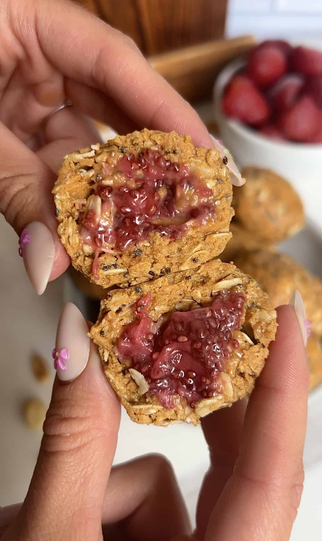 Hands holding a halved PB&J protein energy bite, showing the vibrant strawberry chia jam filling and oat‑peanut butter texture.