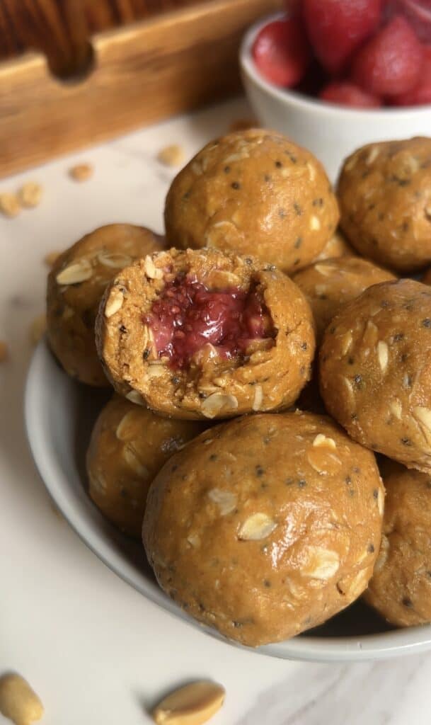 Bowl of PB&J protein energy bites made with oats, peanut butter, and chia seeds, one showing a strawberry jam filling; fresh strawberries in background.