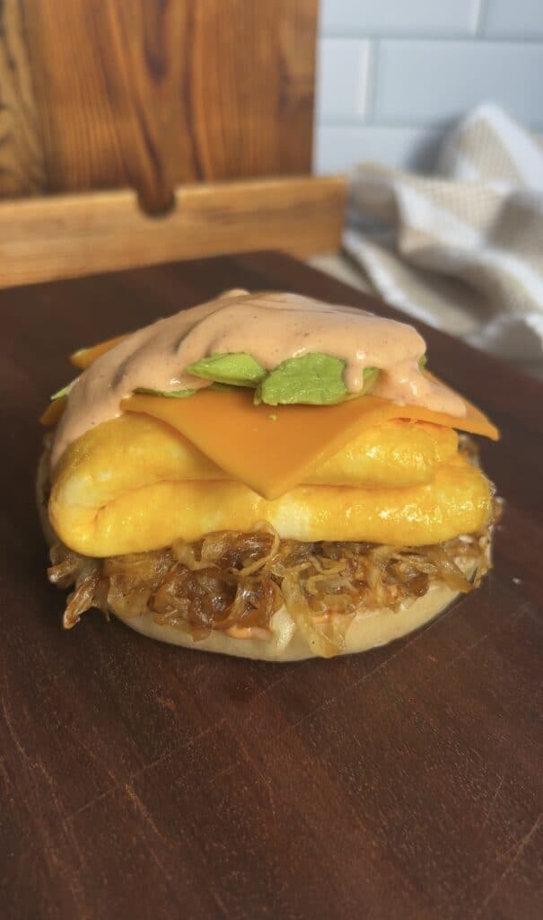 Breakfast bagel topped with creamy eggs, cheddar, avocado, and a generous drizzle of spicy aioli — glossy sauce highlighting rich textures.