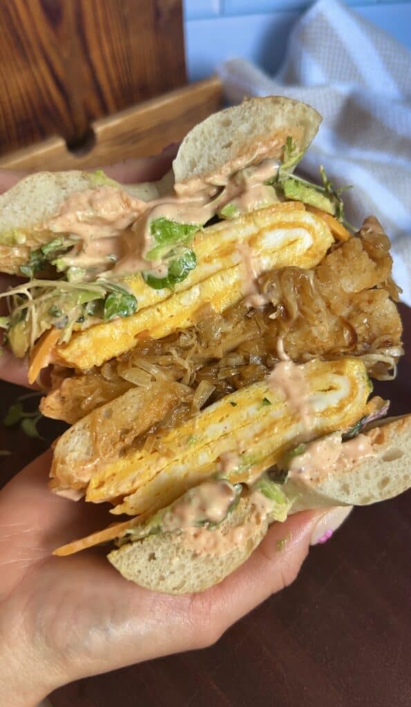Sliced breakfast bagel revealing layers of creamy eggs, caramelized onions, avocado, microgreens, and spicy aioli — perfect for brunch photography.
