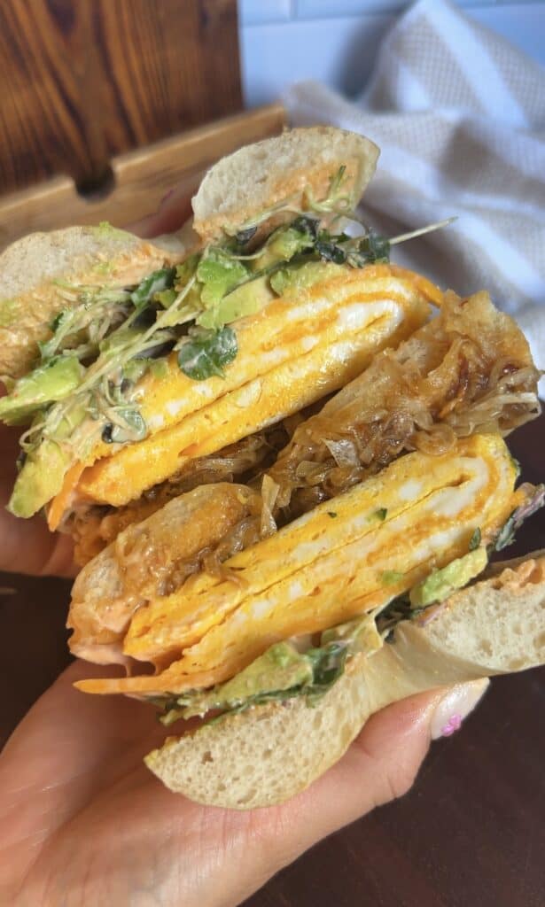 Close‑up of sliced breakfast bagel showing caramelized onions, folded eggs, greens, and spicy aioli — highlighting the sandwich’s rich, savory layers.