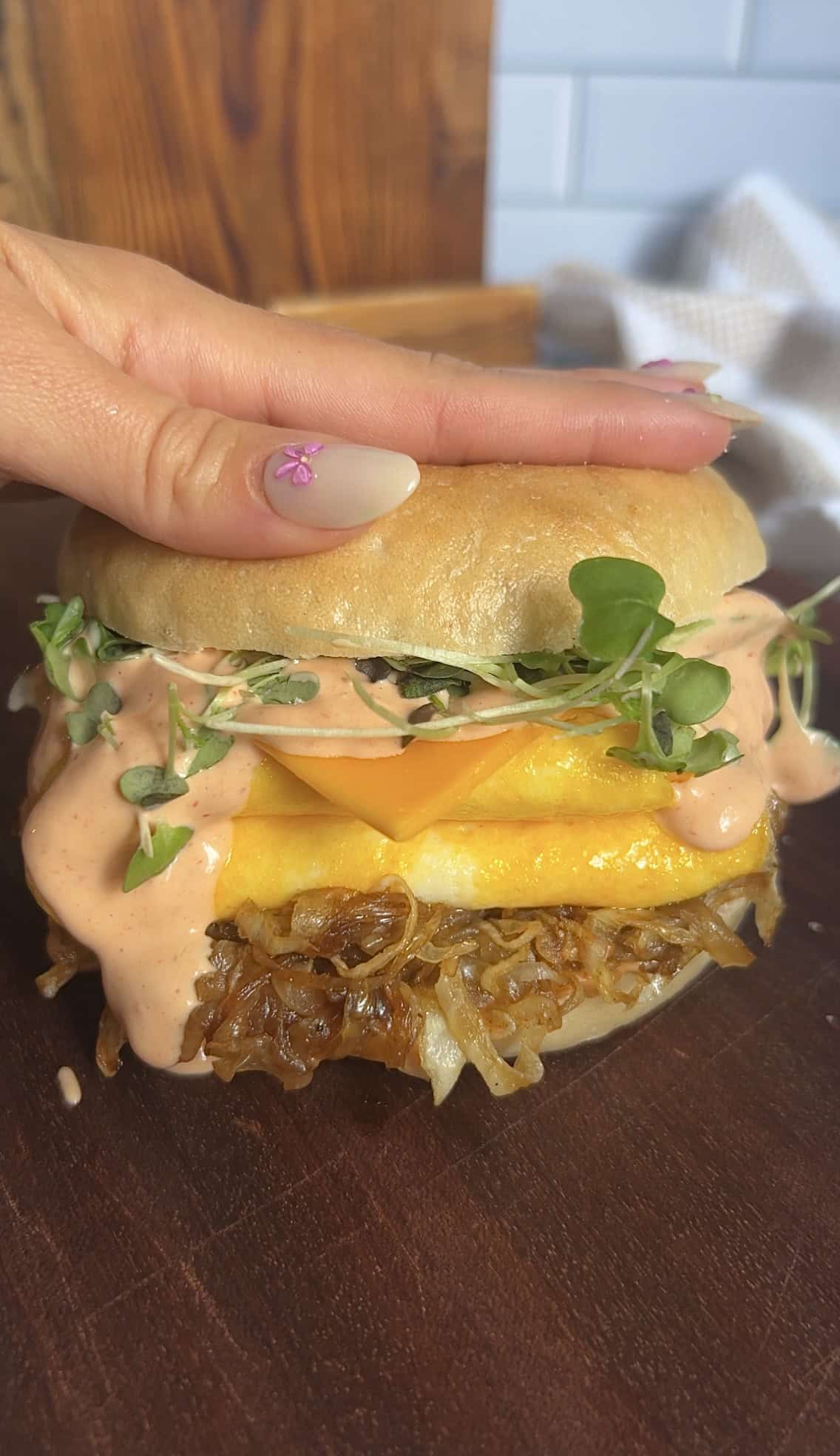 Hand pressing a stacked breakfast bagel with caramelized onions, eggs, cheddar, microgreens, and spicy aioli — creamy sauce dripping for visual appeal.