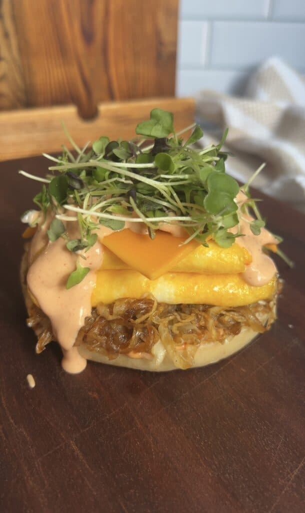Breakfast bagel layered with eggs, cheddar, caramelized onions, spicy aioli, and fresh microgreens — colorful, camera‑ready brunch sandwich.