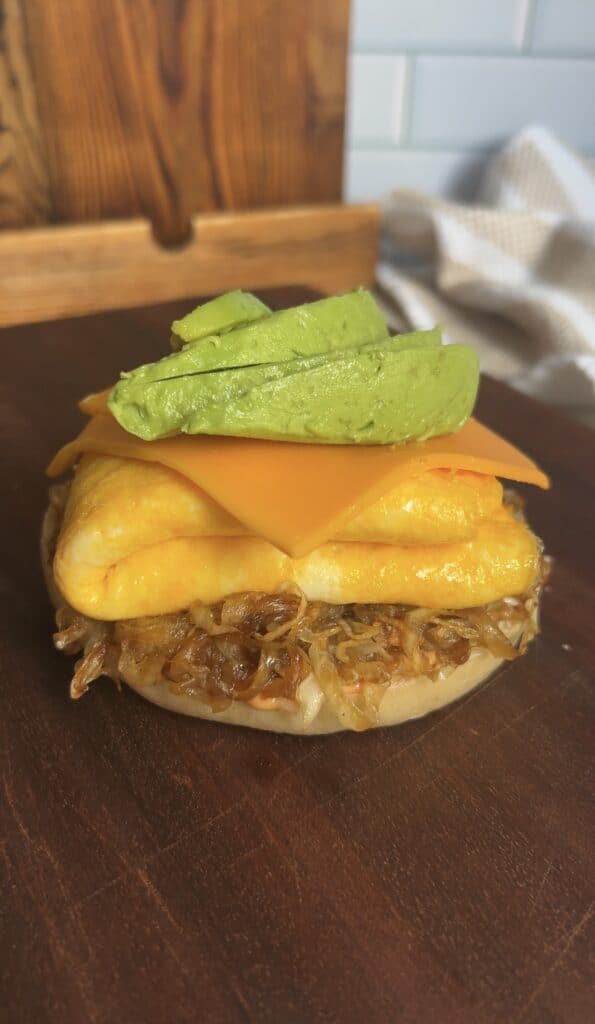 Open‑faced breakfast bagel stacked with caramelized onions, folded eggs, cheddar cheese, and fresh avocado slices — vibrant layers ready for assembly.