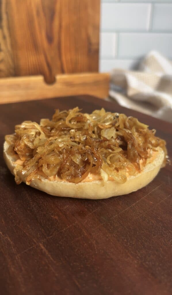 Close‑up of a toasted bagel half topped with golden caramelized onions and spicy aioli, set on a wooden board — the first step in assembling a breakfast bagel sandwich.