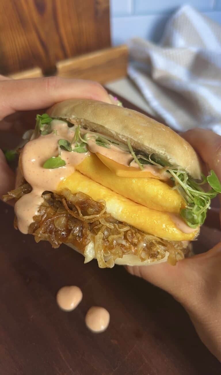 Close‑up of a hand holding a spicy aioli breakfast bagel filled with caramelized onions, creamy eggs, cheddar, and microgreens — sauce dripping down the side.