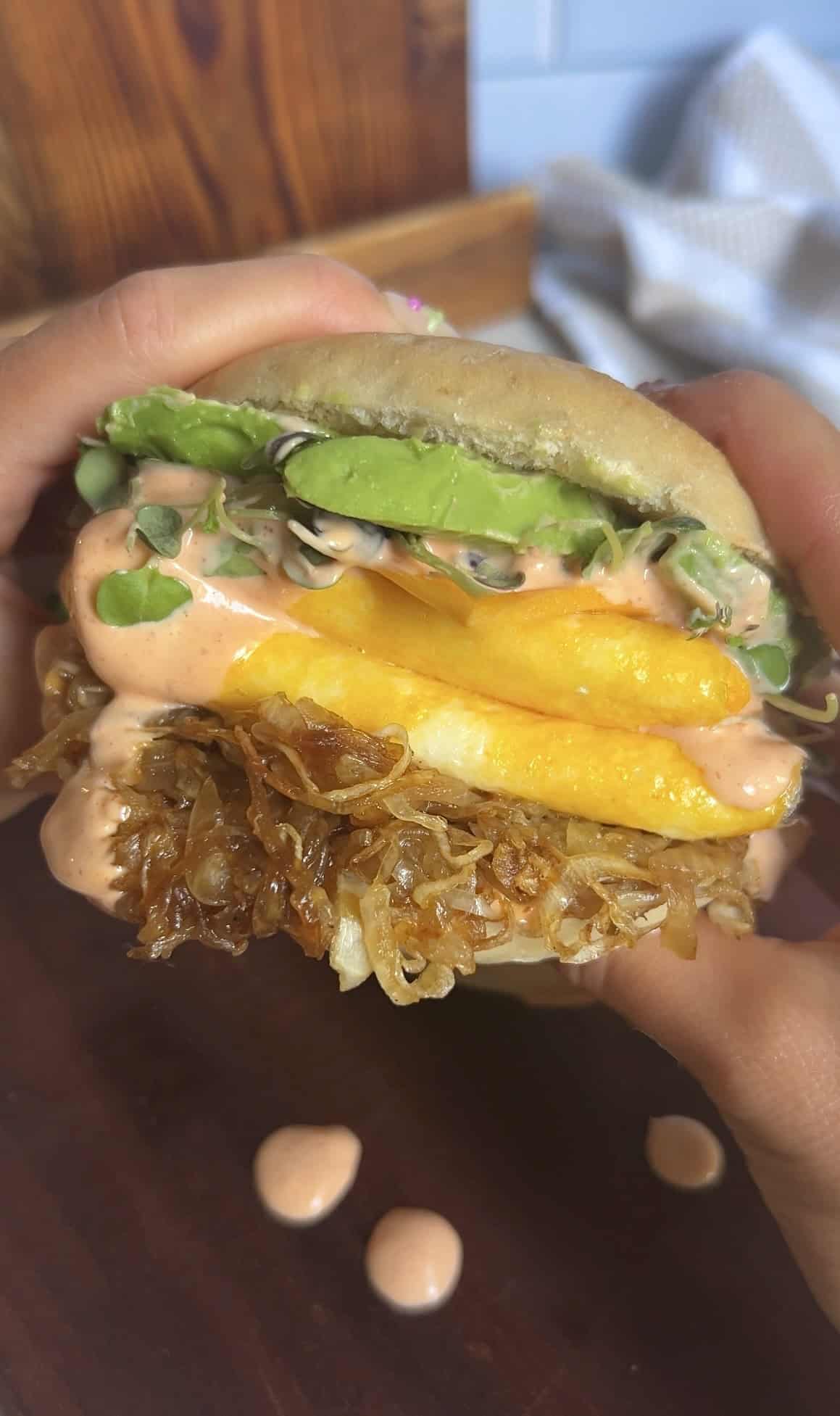 Breakfast bagel sandwich held in hands showing layers of avocado, eggs, caramelized onions, and spicy aioli — rich textures and vibrant colors.